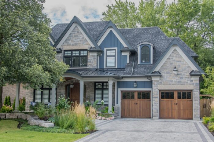 A large modern multi level residential executive home with brown and grey brick stone exterior, shingled roof, beautiful wooden garage doors, interlocked driveway, green gardens and lawn.